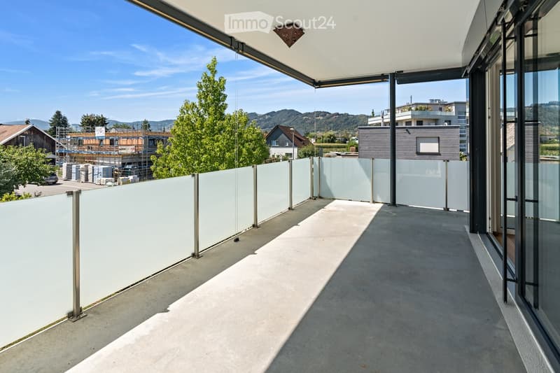 Modern balcony with concrete floor, white glass railing, and metal posts, overlooking a scenic landscape with trees and distant hills.