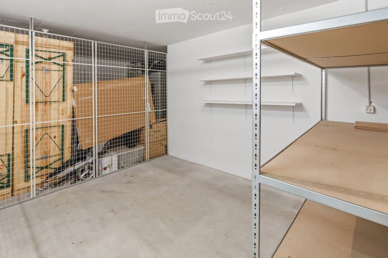 Empty room with white walls, concrete floor, metal shelving unit, and storage area with boxes and cables behind a metal mesh barrier.