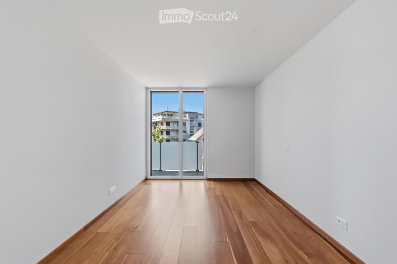 Empty room with light wooden flooring and white walls, featuring a large sliding glass door that opens to a view of buildings and greenery outside.