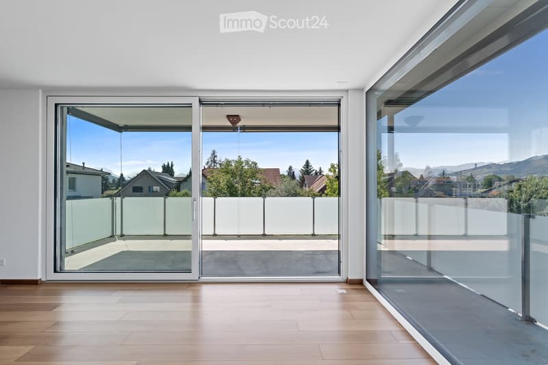 Bright, empty room with large glass sliding doors leading to a balcony, wooden flooring, and white walls.