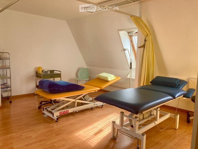 Therapy room with a wooden floor, two adjustable treatment tables, one with a yellow cushion, a green chair, a small desk, and a standing lamp. There is a window with white blinds.