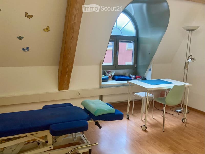Room with blue examination table, two blue chairs, a desk, window seat with a pink building outside, light blue wall decoration of butterflies, white floor lamp