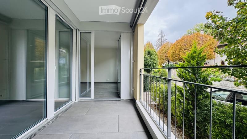 Modern balcony with large glass doors, metal railing, and a view of a green garden and trees.