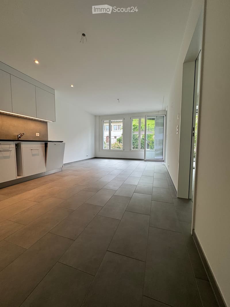 Empty room with tiled floor, white walls, and a small kitchen area with cabinets and a sink.