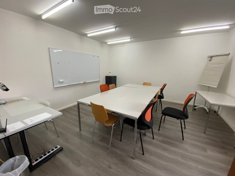 Bright, modern meeting room with white walls, wooden flooring, and fluorescent lighting. Features a large central table surrounded by orange and black chairs, a whiteboard, a projector screen, and a small cabinet.
