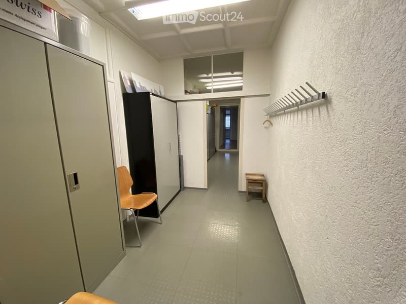 A clean, modern hallway with white walls, tiled floor, and a coat rack on the right side. A small wooden stool and a modern chair are visible, along with a black cabinet and a glass partition leading to another room.