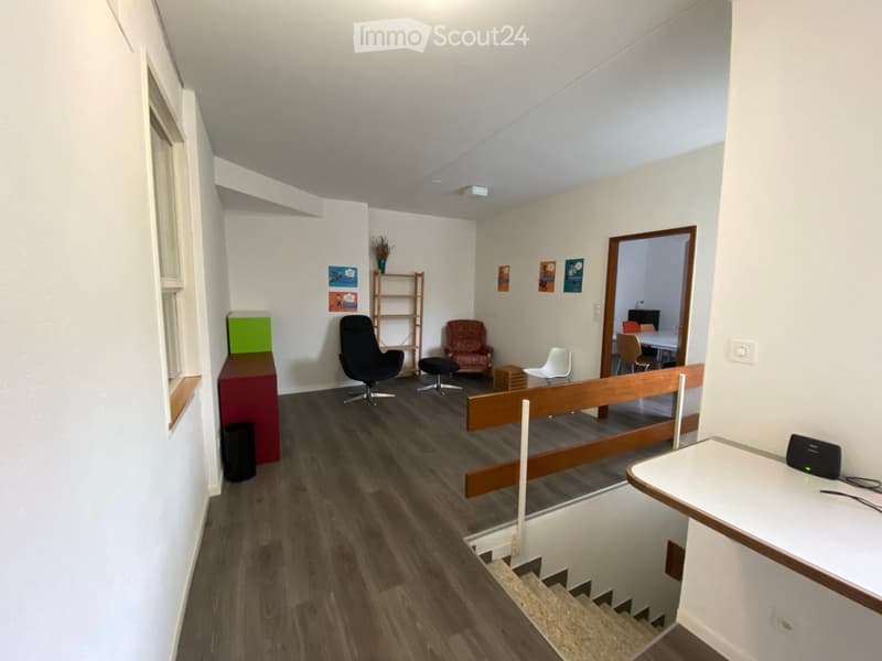 A modern, well-lit room with wooden flooring, white walls, and large windows. The space includes a mix of seating options such as chairs and stools, a small table, a shelf with decorative items, and a staircase leading to another level.