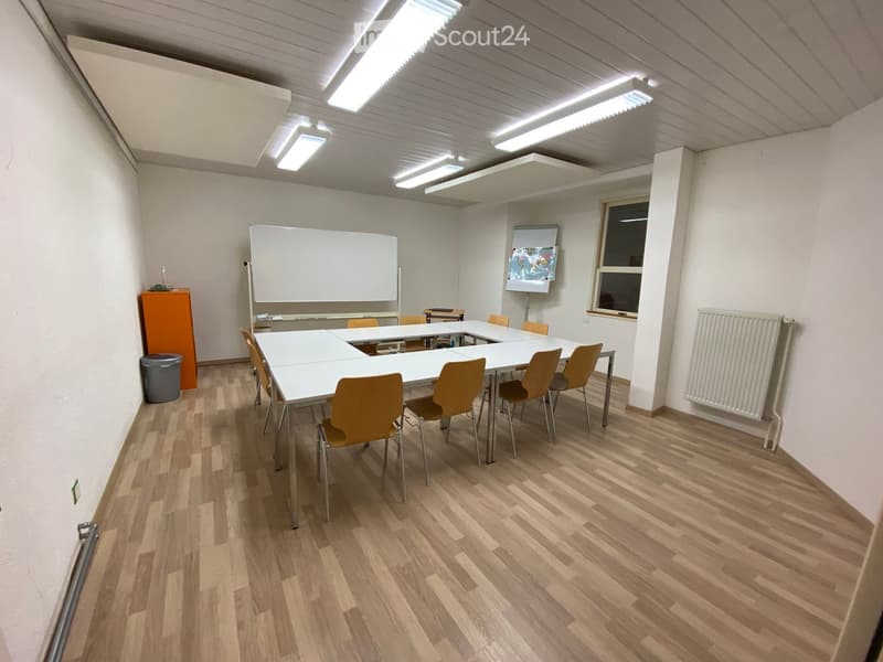 Bright, empty room with wooden flooring, white walls, and a large table surrounded by chairs. Features include a whiteboard, a screen, a radiator, and a small orange cabinet.