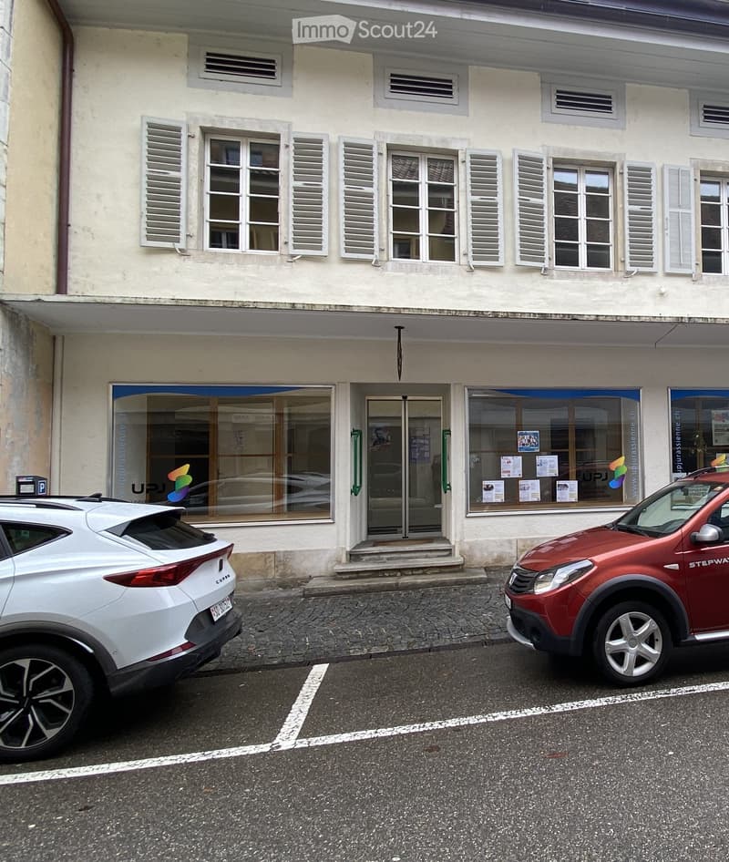 Two-story building with light-colored facade, multiple windows with shutters, and a storefront with glass doors and windows displaying posters and signage. Two cars are parked in front of the building.