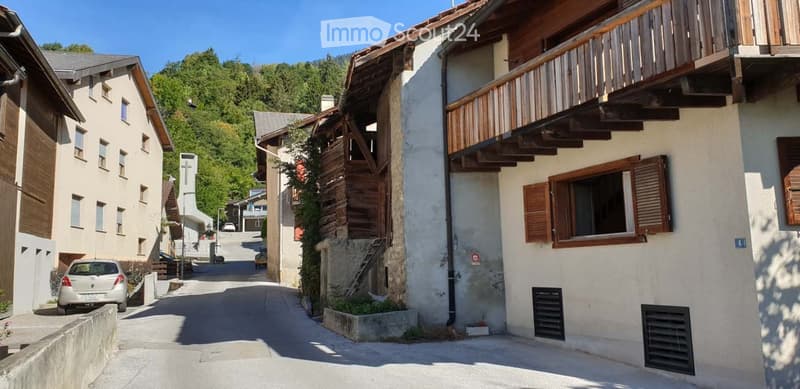 A row of traditional wooden and stone houses in a narrow village street, surrounded by greenery and mountains.
