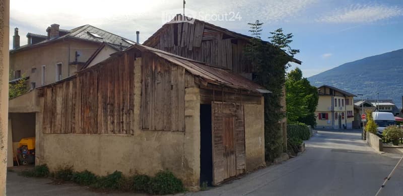 A rustic wooden building with a stone base and a corrugated metal roof, situated on a narrow street with other residential structures and a mountainous backdrop.