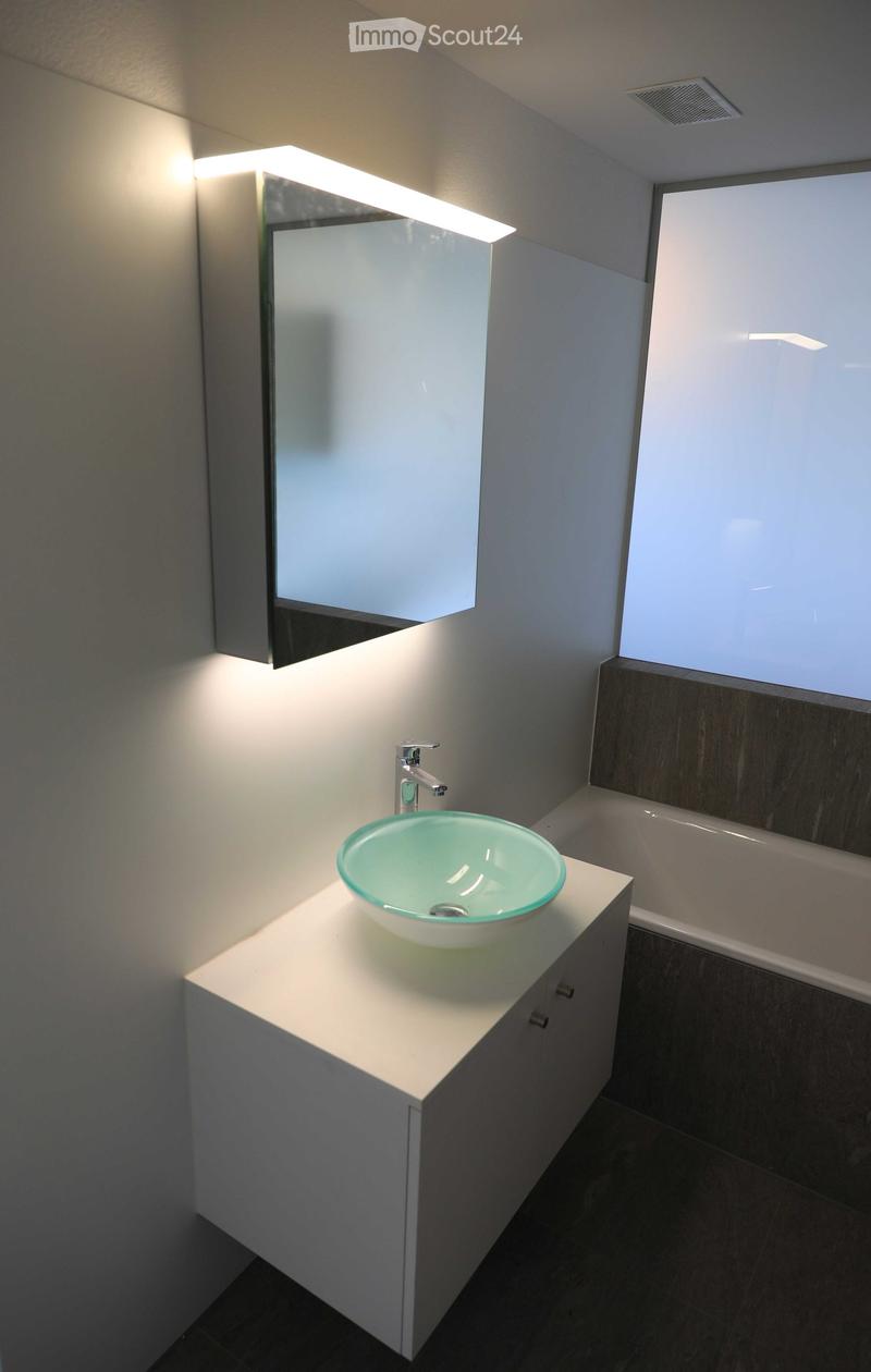Modern bathroom with a white vanity, a green glass sink bowl, a sleek faucet, a large mirror with built-in lighting, and a bathtub visible in the background.