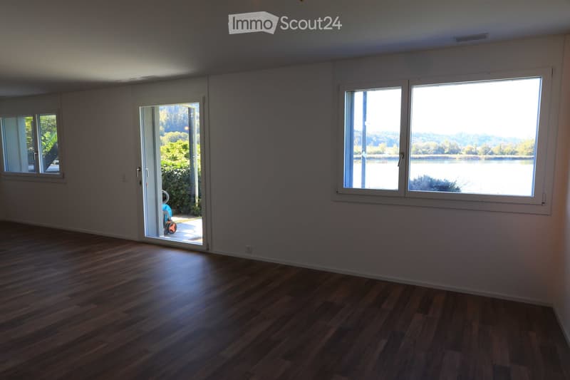 Empty room with wooden flooring, large windows offering a scenic view of a lake and surrounding greenery, and a sliding glass door leading outside.