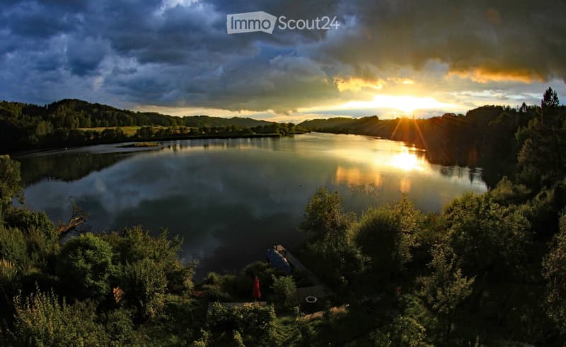 A serene lakeside view at sunset, with calm water reflecting the golden light, surrounded by lush greenery and trees.
