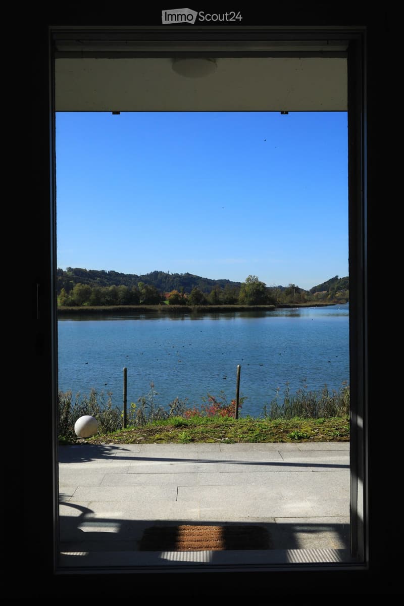 View from a large window or balcony overlooking a serene lake with a clear blue sky, distant hills, and a paved patio area with a decorative sphere.