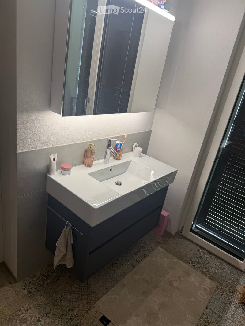 Modern bathroom with a white double sink, chrome faucet, large mirror, and various toiletries on the counter. A towel hangs from a cabinet below the sink.