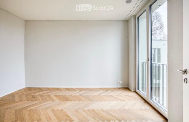 Empty room with light-colored wooden flooring and white walls, featuring a large glass sliding door leading to a balcony.