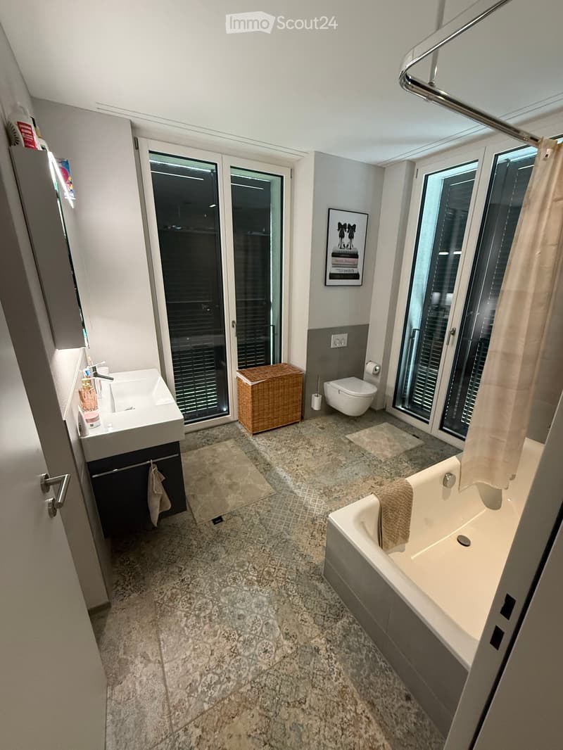 Modern bathroom with a sink, bathtub, toilet, wicker basket, and a picture frame on the wall. The space features large windows and a shower curtain.