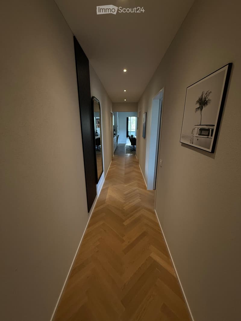 A narrow hallway with herringbone-patterned wooden flooring, beige walls, and recessed lighting. A large mirror and framed artwork are visible on the walls, with a glimpse of an adjacent room featuring a desk and chairs.