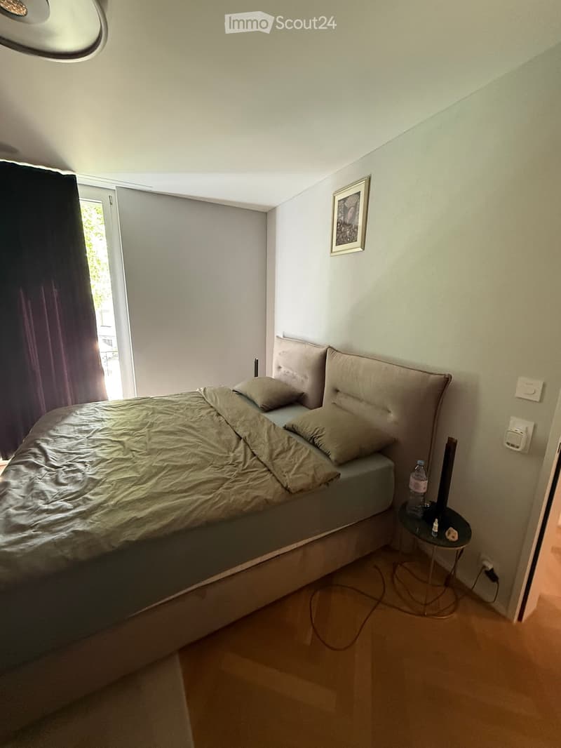 A small bedroom with a single bed, pillows, a nightstand with a bottle, and a window with curtains.