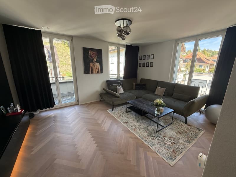 Modern living room with wooden flooring, large windows, grey sectional sofa, coffee table, and decorative rug.
