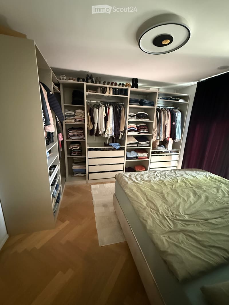 A well-organized bedroom with a neatly made bed, a large wardrobe with multiple sections for hanging clothes and storing folded items, and several drawers for additional storage. The room features wooden flooring and a modern ceiling light.