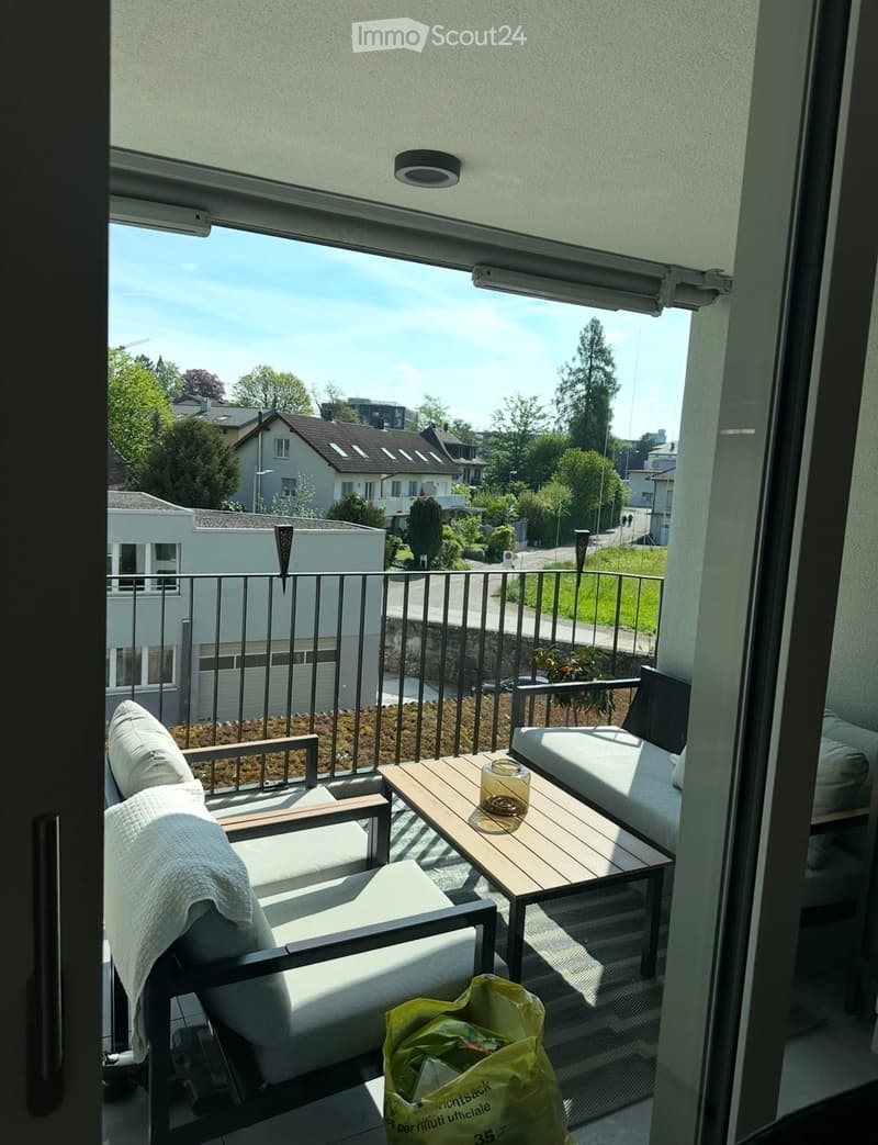 Balcony with a sofa, table, and chair, overlooking a residential area with houses and greenery.