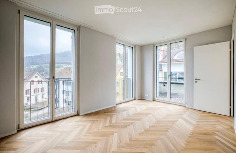 Empty room with wooden herringbone flooring, large windows, and white walls. No furniture or equipment present.