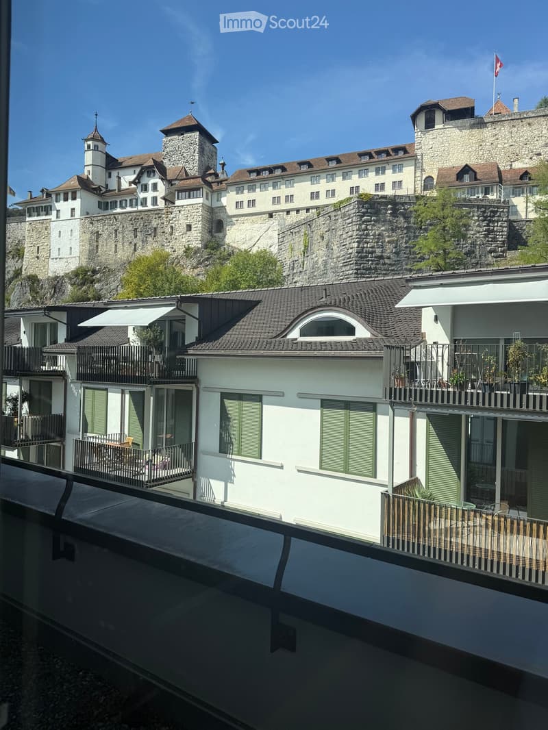 View of a historic castle and modern buildings with balconies and outdoor furniture, overlooking a scenic cityscape.