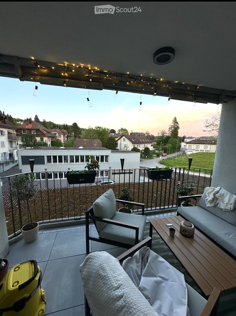 Balcony with a view of the city, featuring a sofa, chair, small table, potted plants, and string lights.