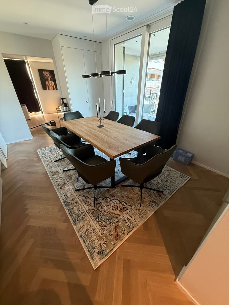 Dining area with a wooden dining table, six black chairs, a patterned rug, and a candle holder on the table. The room has wooden flooring and large windows with dark curtains.