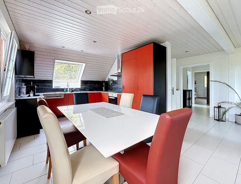 Modern kitchen and dining area with white dining table, red and beige chairs, red and black cabinets, and stainless steel appliances.