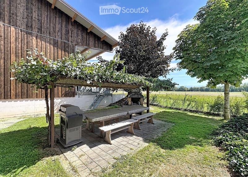Outdoor patio area with a wooden pergola, stone benches, a barbecue, and a brick barbecue pit, surrounded by trees and a grassy lawn.