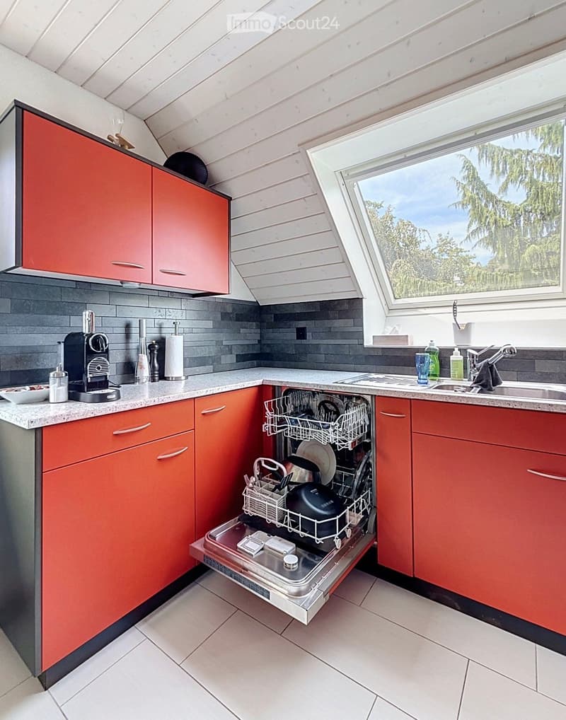 Modern kitchen with red cabinets, white tiled floor, and a dishwasher open, showing dishes inside. There is a coffee maker on the counter, a sink with a faucet, and various small items like cups and bottles.