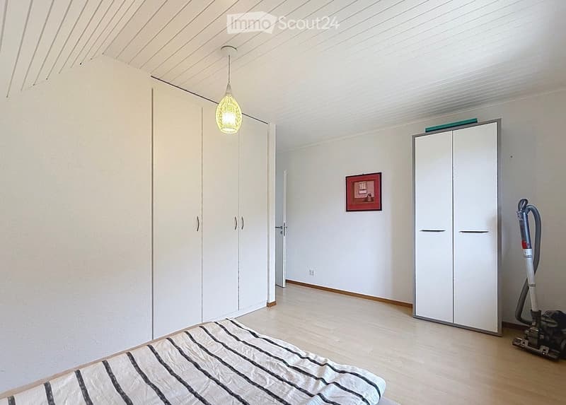 Bright, minimalist bedroom with white walls, wooden flooring, and modern built-in wardrobes. Features a single bed with a striped duvet, a hanging pendant light, and a vacuum cleaner in the corner.