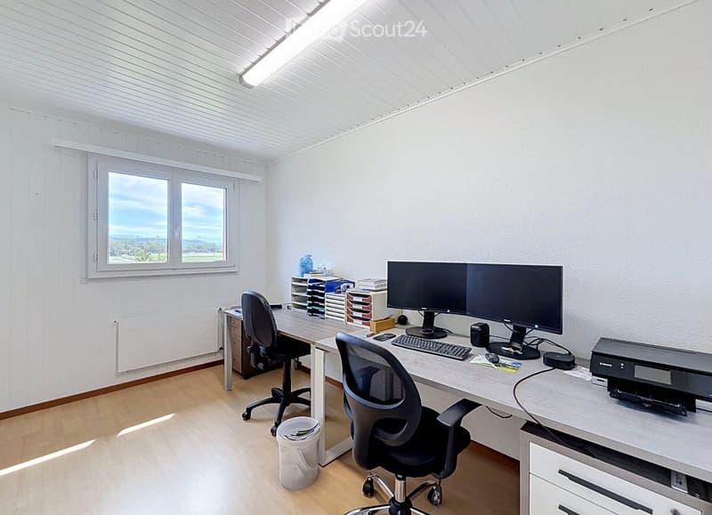 Bright, modern office space with a large window offering a scenic view, featuring a desk with dual monitors, a keyboard, a mouse, a printer, and two ergonomic chairs.