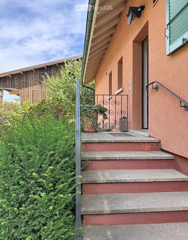 Entrance to a two-story house with beige walls, concrete steps, metal railing, and small garden area with plants and a lantern.