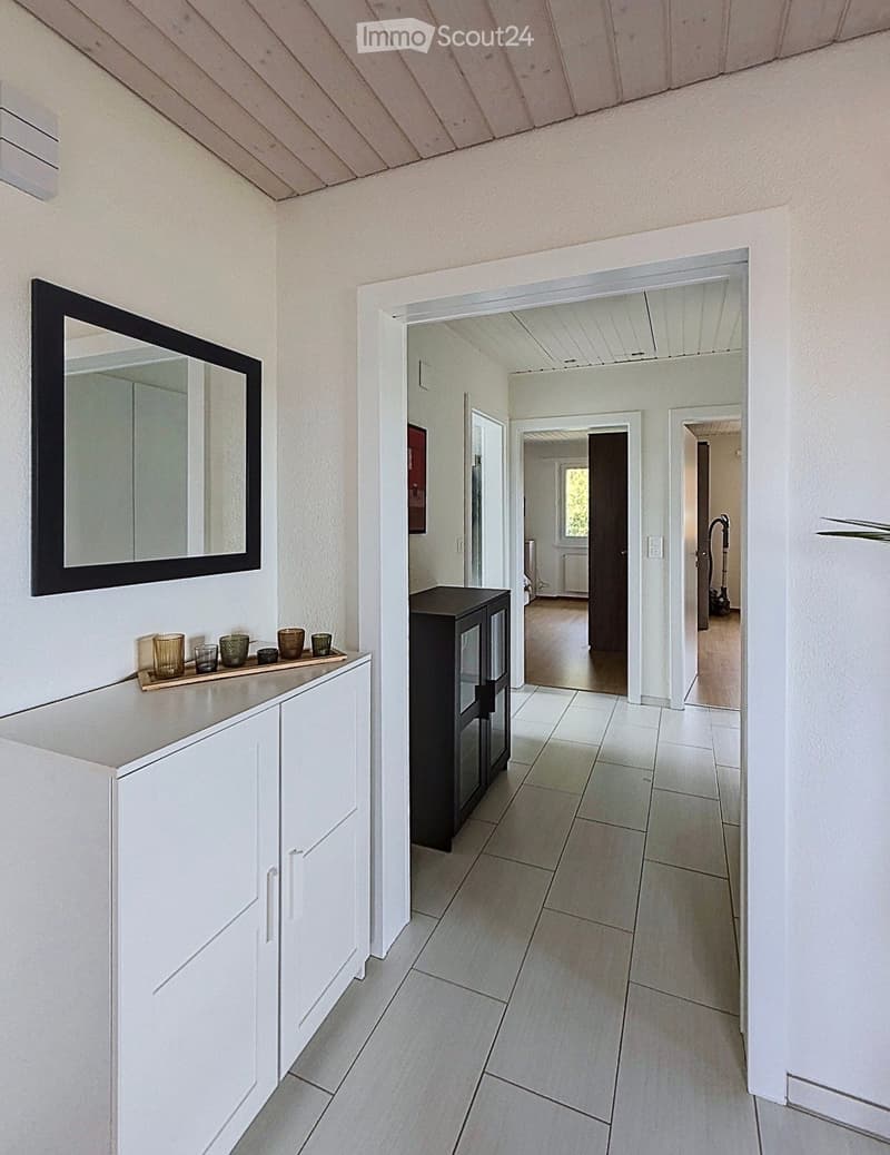 Bright hallway with white tiled flooring, white walls, and a slanted wooden ceiling. Features a small white cabinet with a mirror, a black glass-fronted cabinet, and several small decorative items on a shelf.