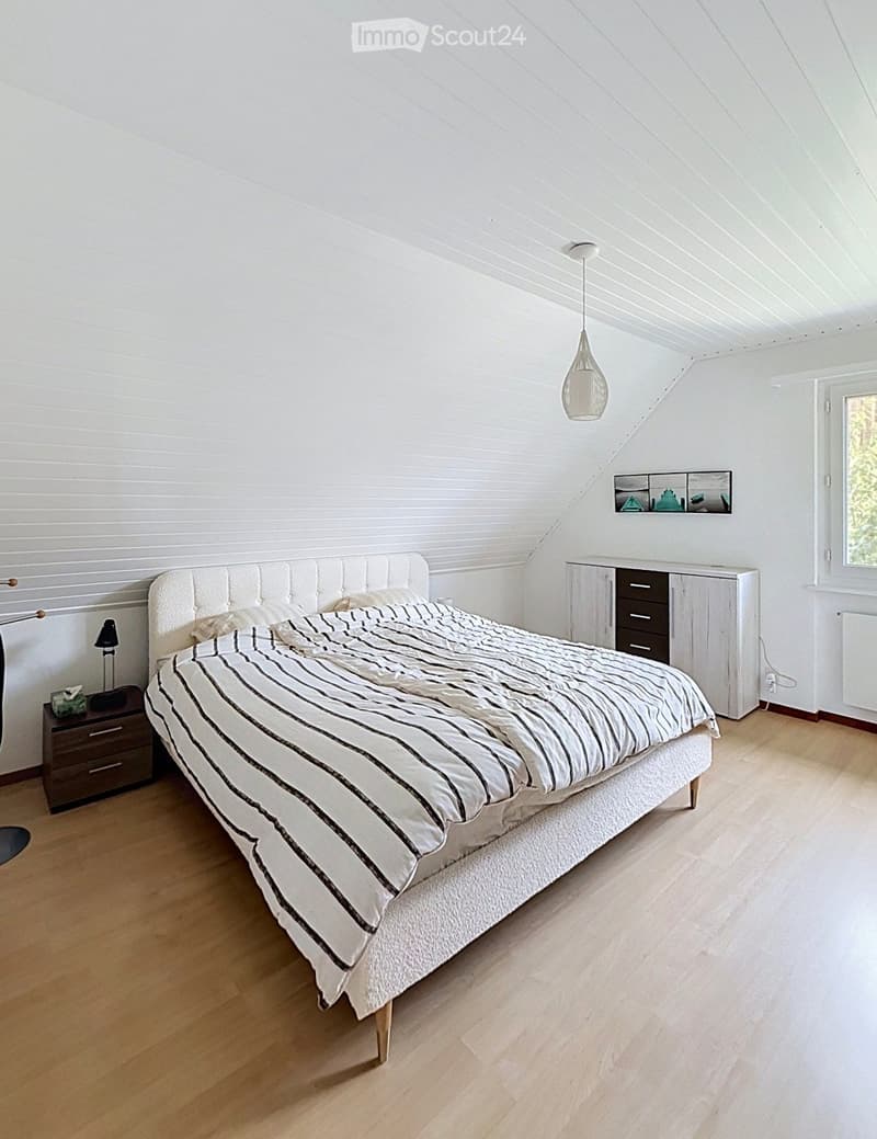 Bright bedroom with wooden flooring, white walls, and a slanted ceiling. Features a large bed with striped bedding, a nightstand with a lamp, a white cabinet with drawers, and a wall-mounted mirror.