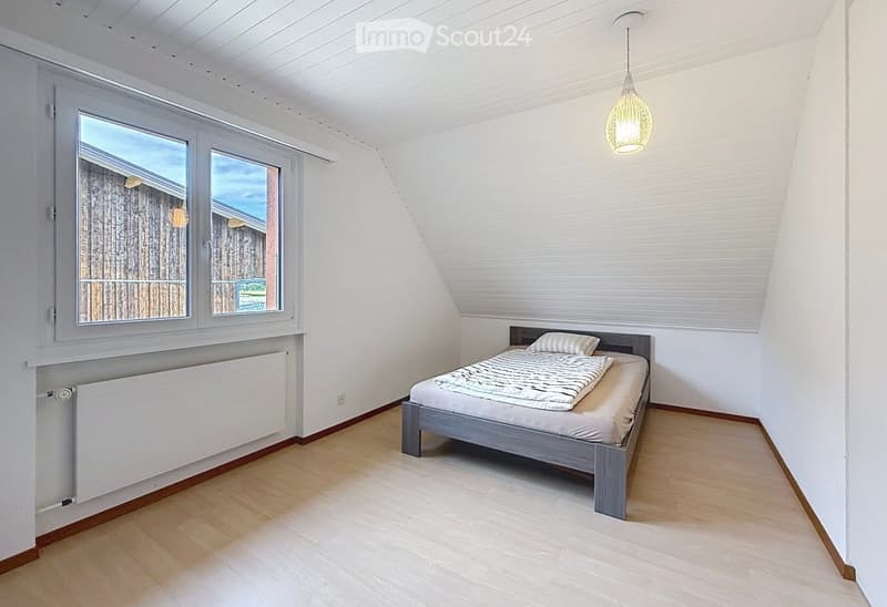 Bright bedroom with wooden flooring, white walls, and a slanted ceiling. Features a single bed with a grey frame and bedding, a radiator below a large window, and a hanging pendant light.