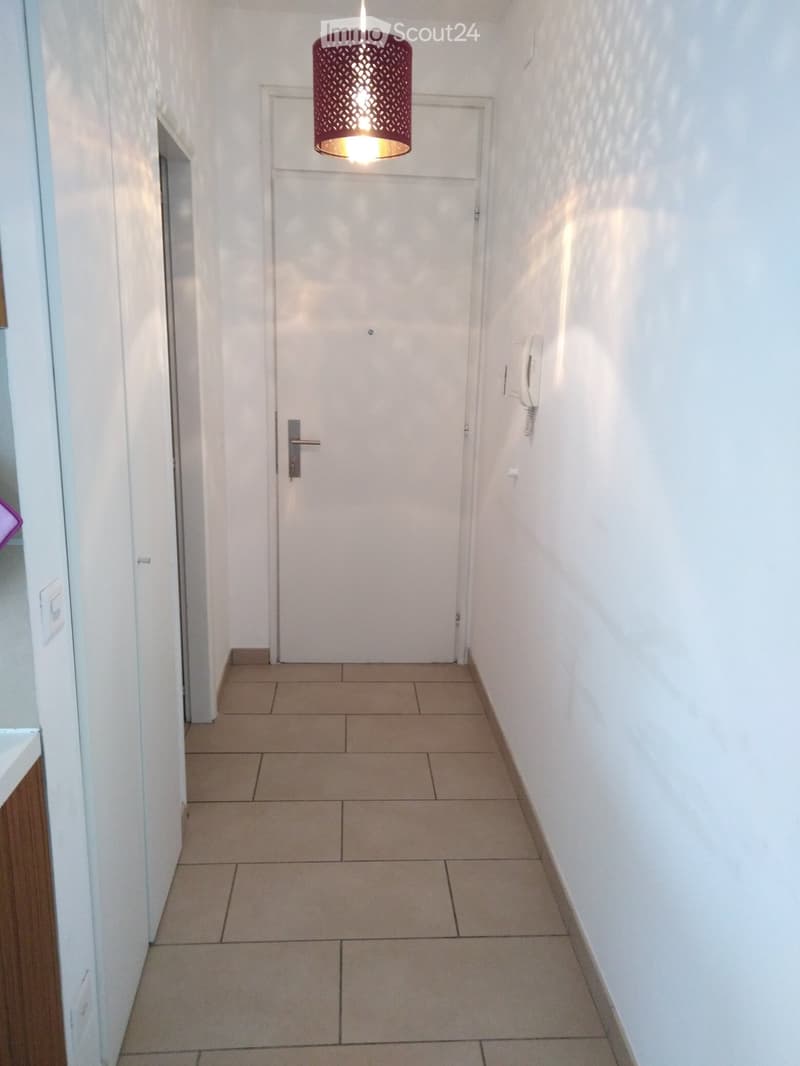 A narrow hallway with tiled flooring, white walls, and a single door at the end. A hanging light fixture with a red shade illuminates the space.