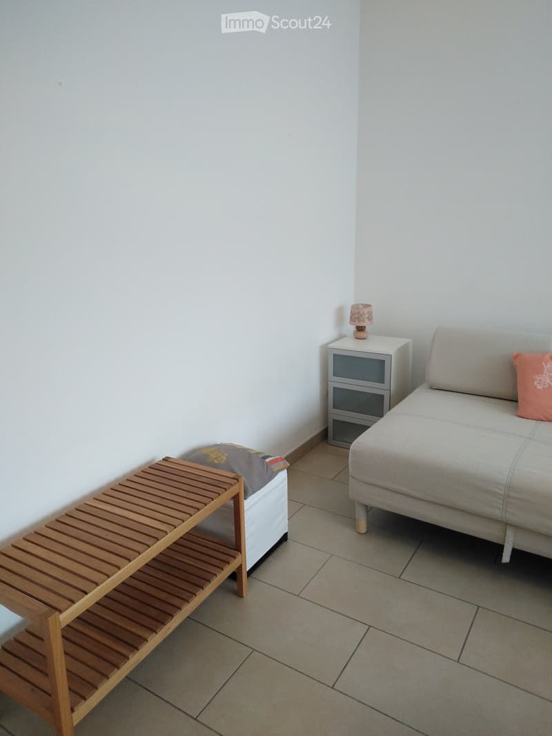 A small, modern living room with a grey sofa, wooden bench, nightstand, and lamp.