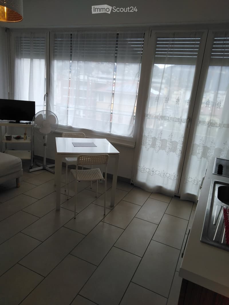 A small, well-lit room with tiled flooring, a white table with two chairs, a standing fan, a television on a small stand, and a cup on the stand.