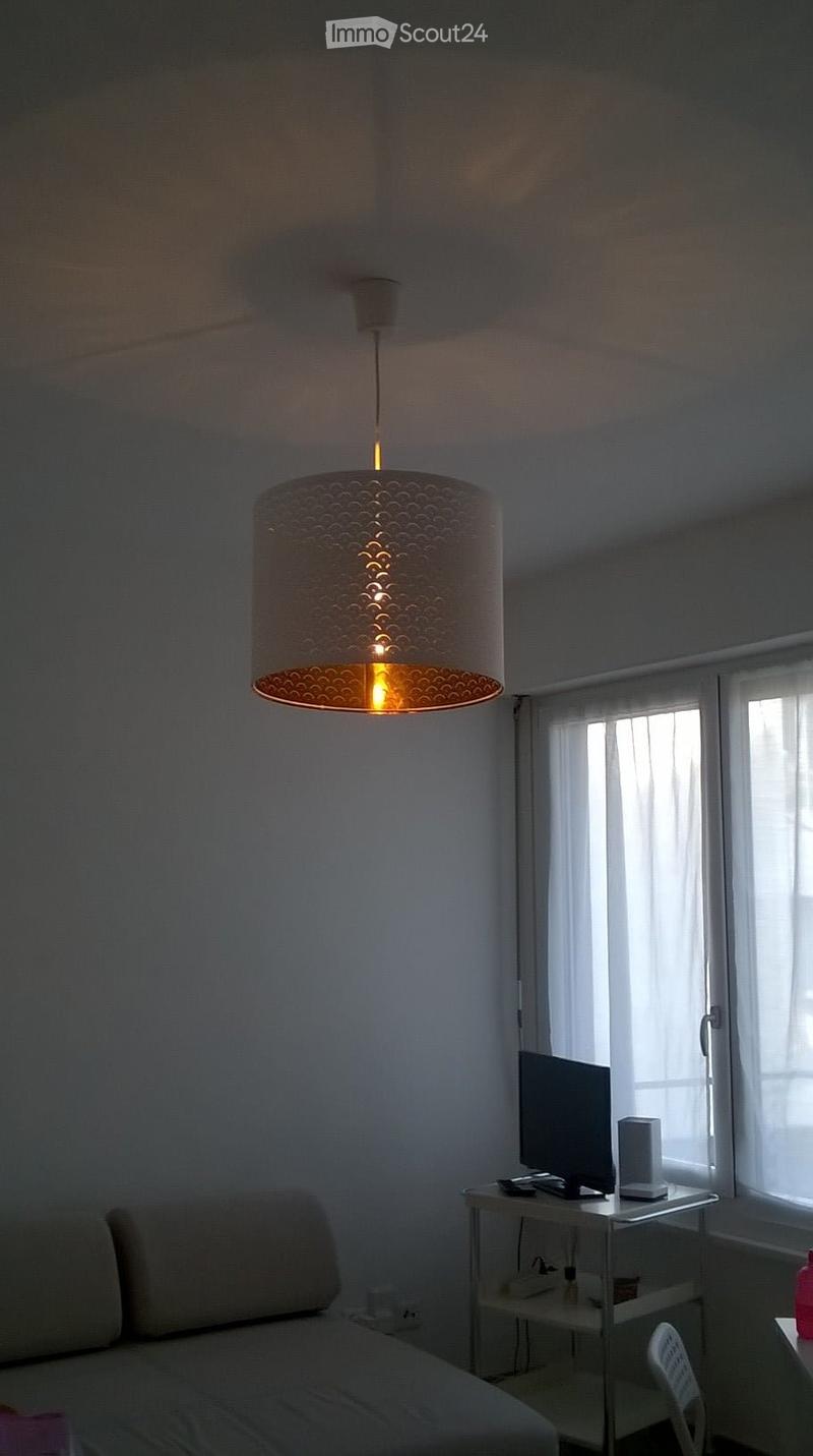 A small, modern bedroom with a bed, a small table with a screen and lamp, and a hanging decorative light fixture.