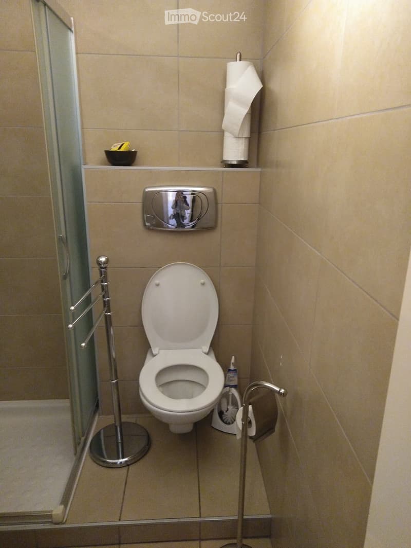A small bathroom with beige tiled walls and floor, featuring a toilet, a towel rack, a toilet paper holder, a toilet brush, a mirror, and a small bowl on the shelf.