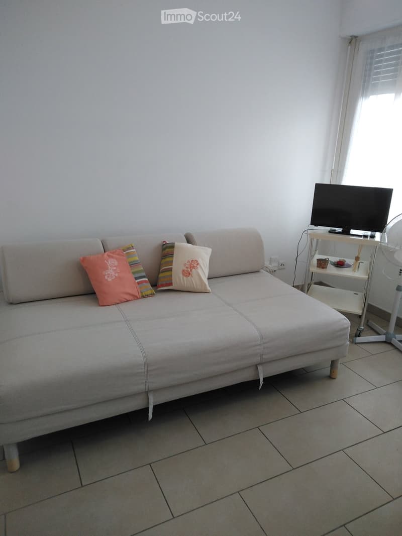 A modern living room with a grey sofa, colorful cushions, a small table with a TV, and a fan near the window.