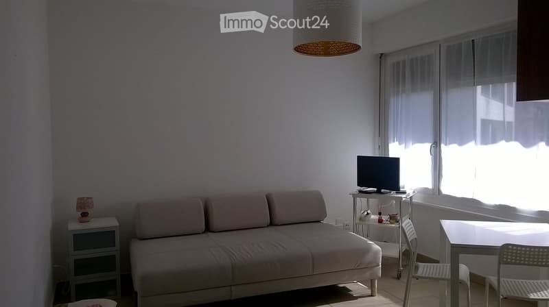 A modern living room with a beige sofa, a small table with a television, a nightstand with a lamp, and a dining area with a table and chairs.