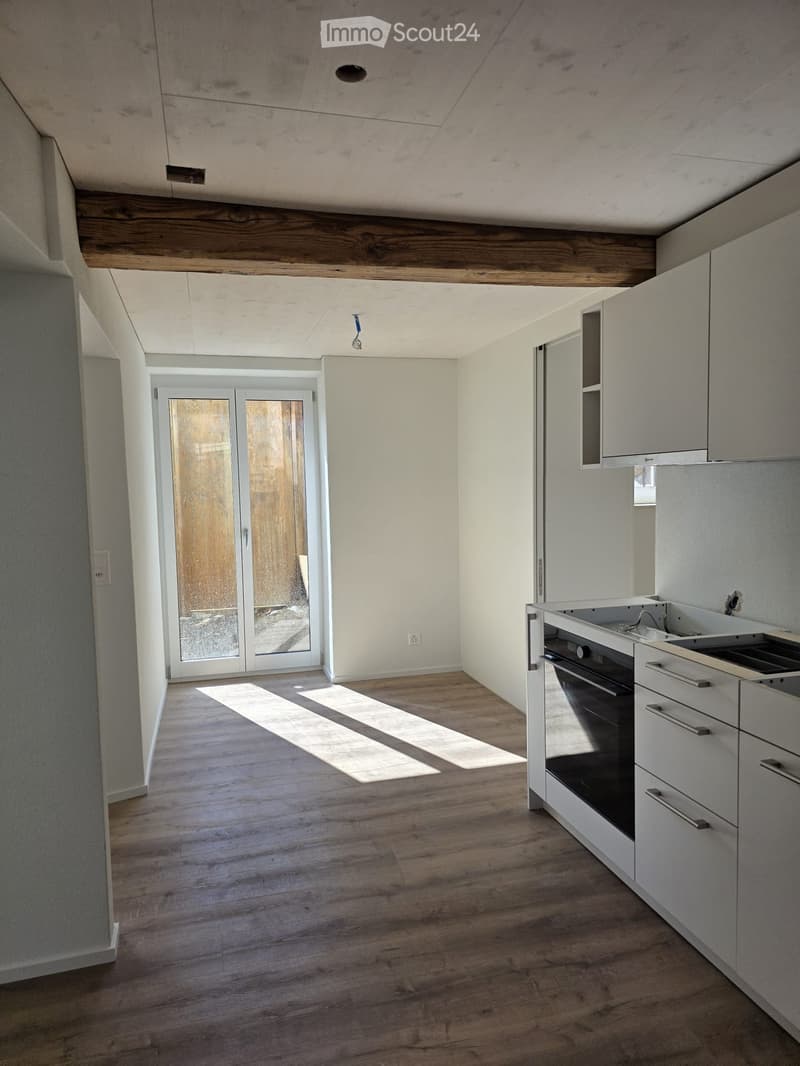 Empty kitchen with wooden flooring, white cabinets, and a built-in oven. Sunlight streams in from a glass door leading to an outdoor area.