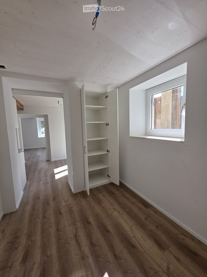 Empty room with wooden flooring, white walls, and a built-in cabinet with shelves. A window on the right side allows natural light to enter.