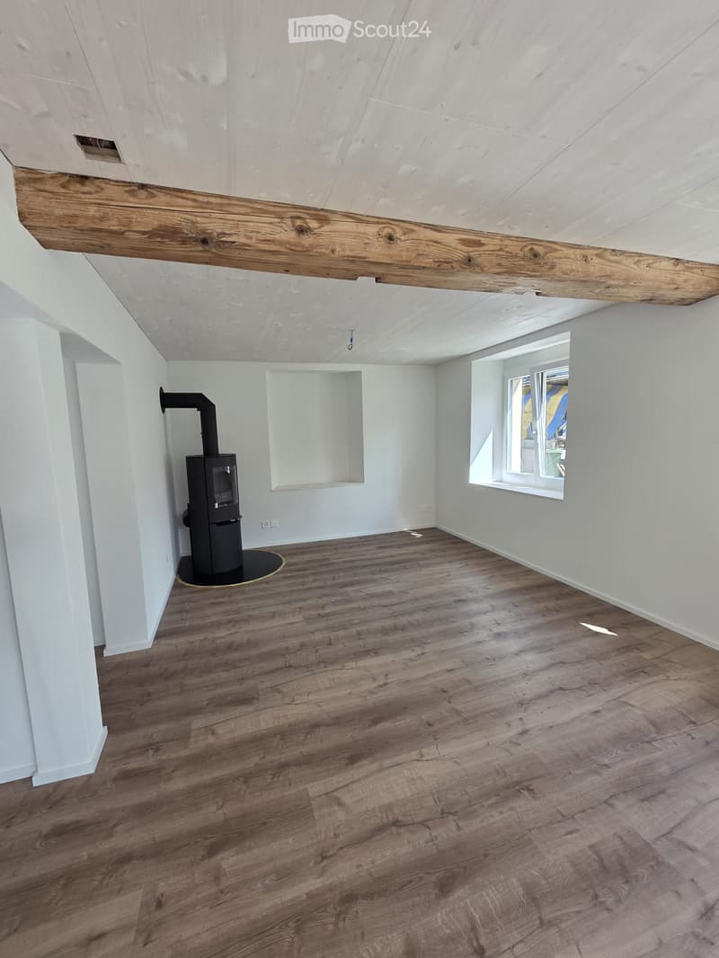 Empty room with wooden flooring, white walls, exposed wooden beam, and a black wood-burning stove with a chimney pipe.